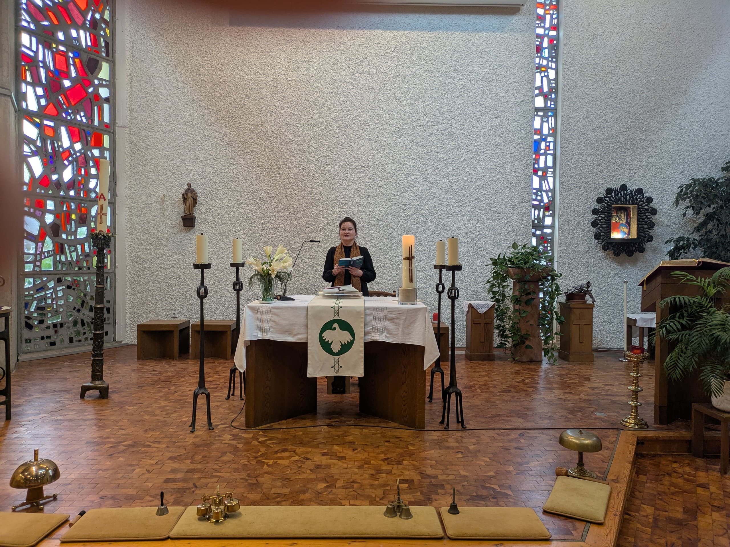 In een kerk met kleurrijke glas-in-loodramen staat een vrouw achter een altaar. Ze houdt een boek vast en kijkt naar de congregatie. Op het altaar staan kaarsen en bloemen.