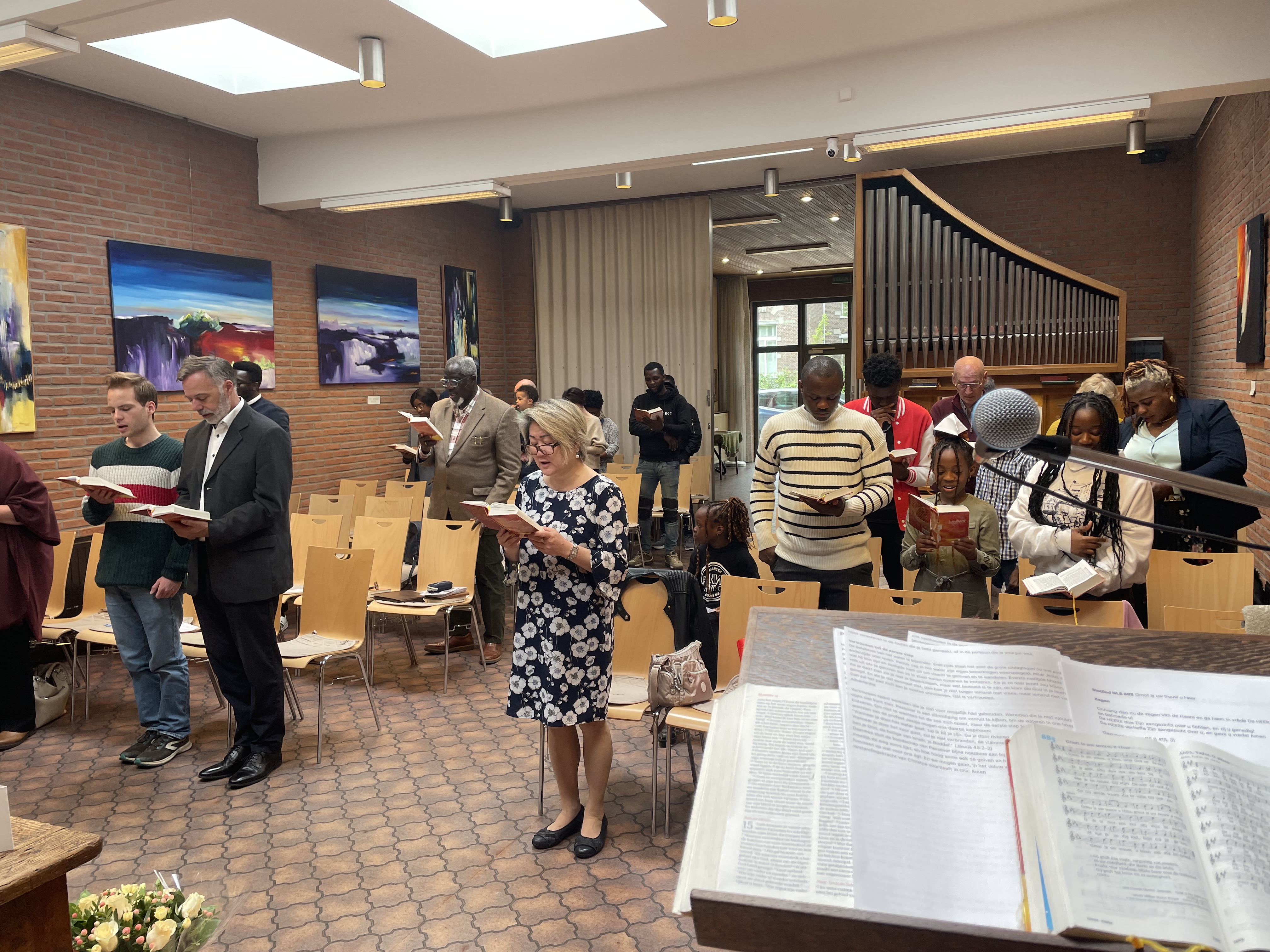 Een groep mensen staat in een kerk, zingend met gezangboeken in hun handen, omringd door houten stoelen en kleurrijke kunst aan de muren. Op de voorgrond is een muziekstand met bladmuziek zichtbaar.